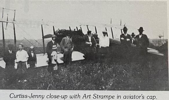A vintage black-and-white photo of people in early 20th-century attire standing in front of a Curtiss Jenny biplane.
