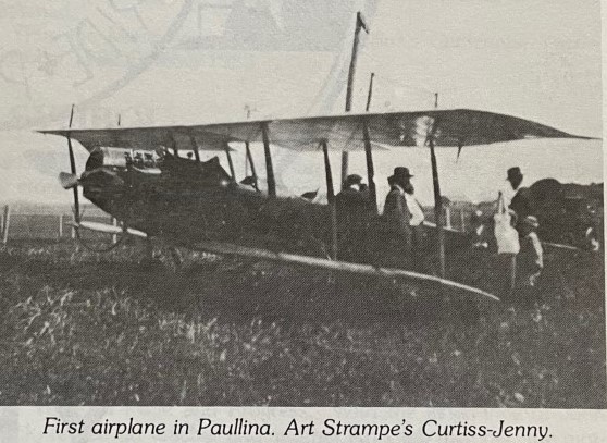 Historic black-and-white photo of a Curtiss-Jenny biplane on grassy field, with several people gathered around.