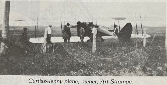Black and white photo of a Curtiss-Jenny biplane with several people gathered around it in a field.