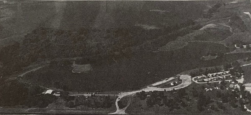 Aerial view in grayscale shows a rural landscape with dense woods, open fields, and a small residential area.