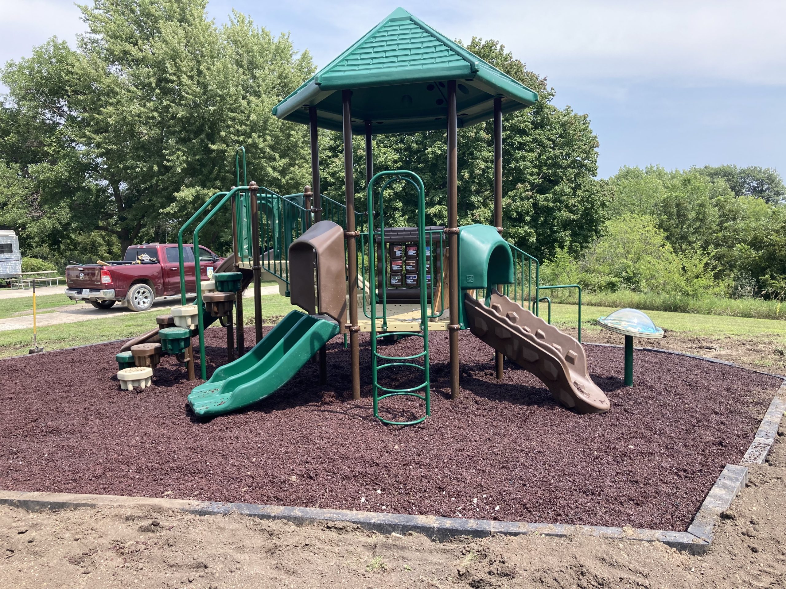 Playground with green and brown climbing structures, slides, and tunnel under a sunny blue sky.