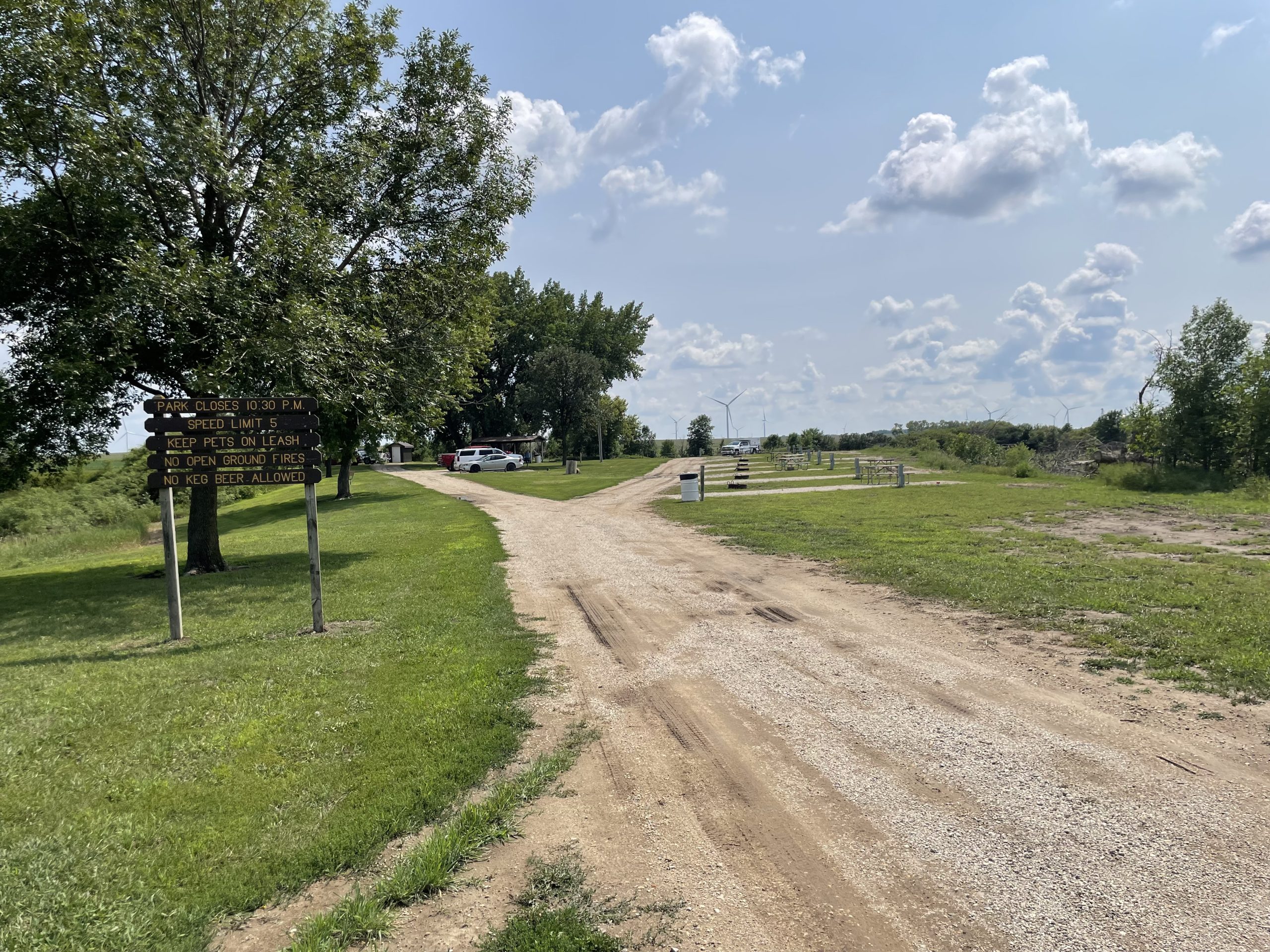 Gravel road leading through an empty campground with picnic tables and fire rings.