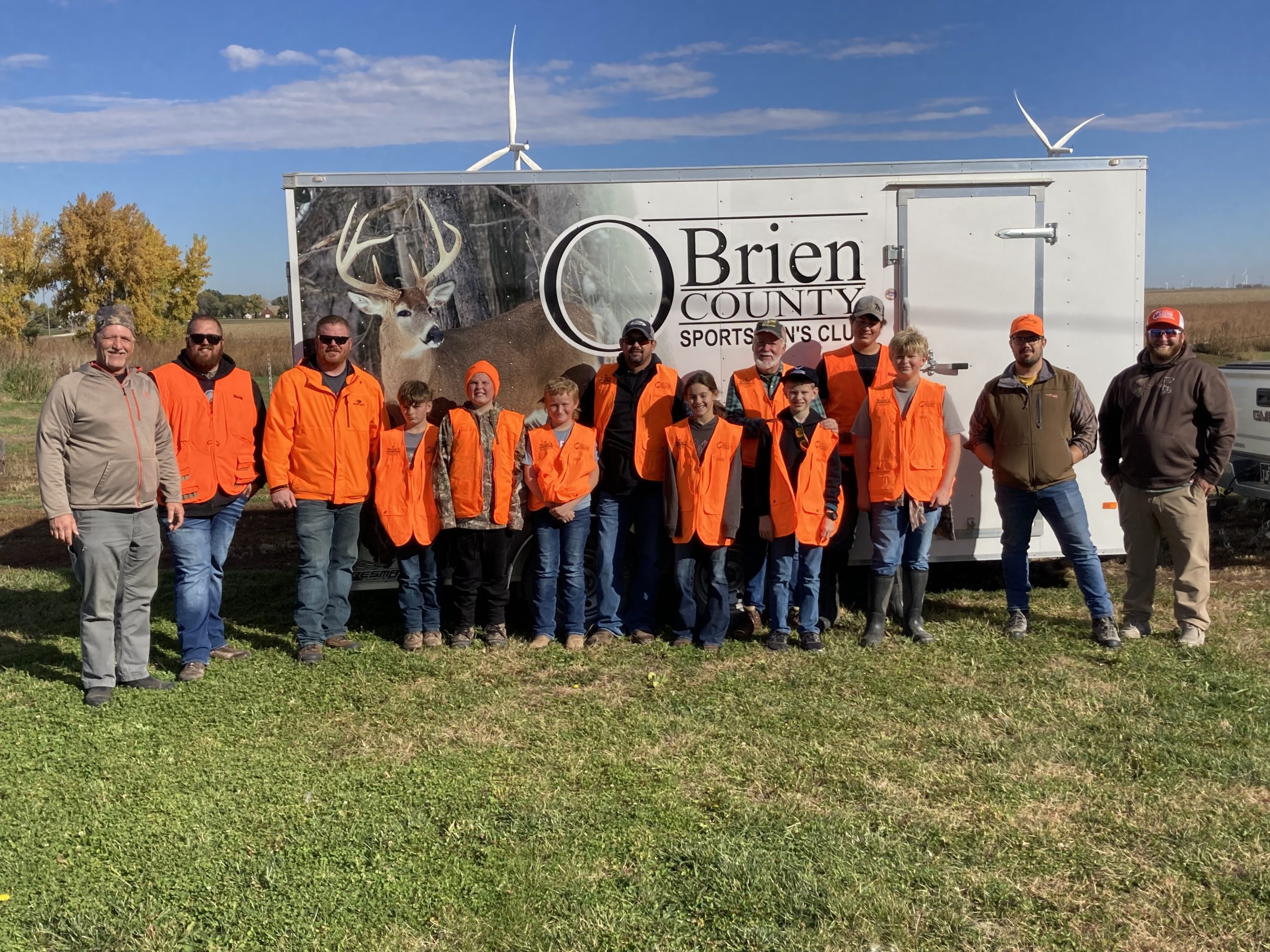A group of 13 people in bright orange vests and jackets stand smiling.