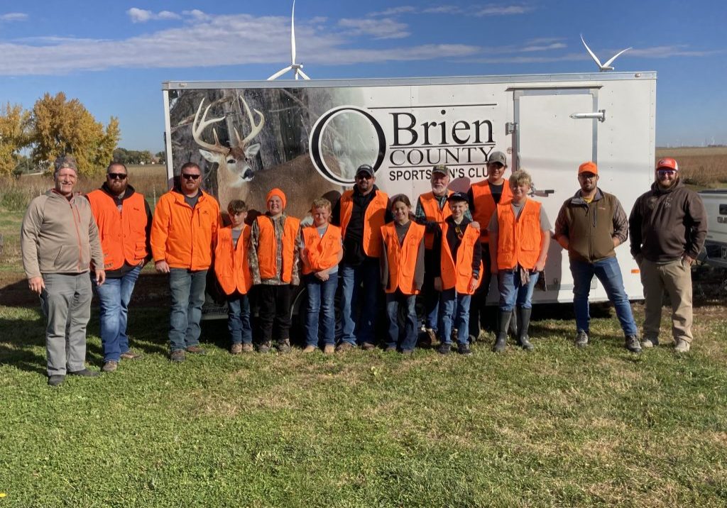 A group of 13 people in bright orange vests and jackets stand smiling.