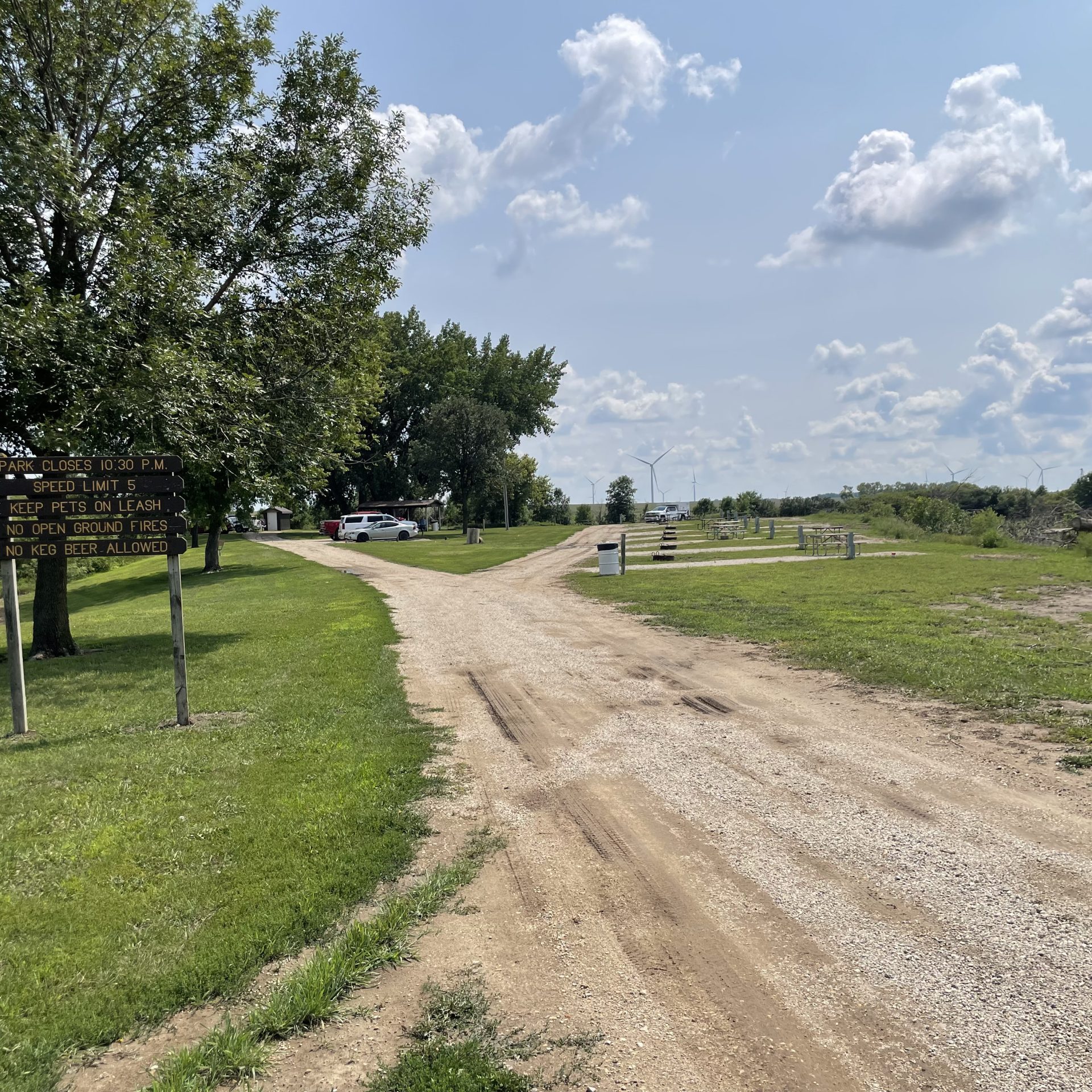 Gravel road leading through an empty campground with picnic tables and fire rings.