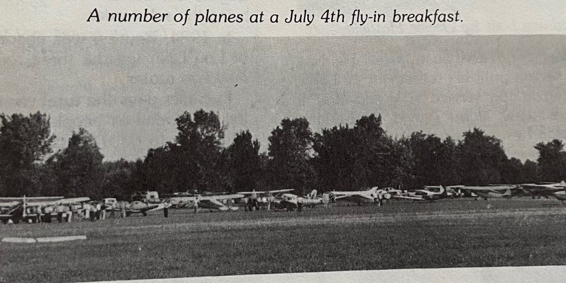 Several small airplanes lined up on a grassy field with trees in the background during a July 4th fly-in breakfast event.